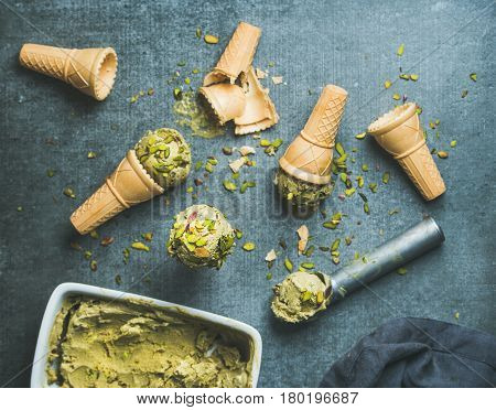Homemade pistachio ice cream in ceramic mold and metal scooper with crashed pistachio nuts and waffle cones over grey concrete background, top view, selective focus