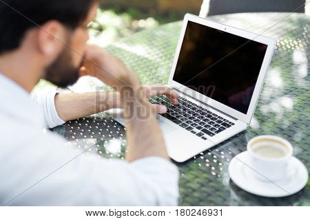 Bearded financial manager with rolled up shirt sleeves searching mistake in his calculations on laptop while drinking coffee in outdoor cafe, over shoulder view