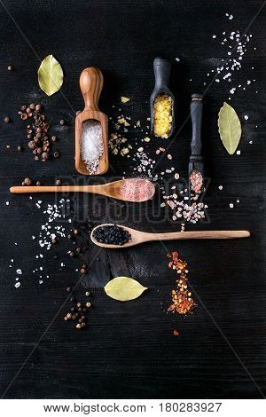 Variety of different colorful salt yellow saffron, pink, black himalayan, white sea and fleur de sel in wooden spoons with black, chili, allspice pepper over black burnt wood background. Top view