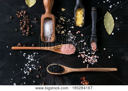 Variety of different colorful salt yellow saffron, pink, black himalayan, white sea and fleur de sel in wooden spoons with black, chili, allspice pepper over black burnt wood background. Top view