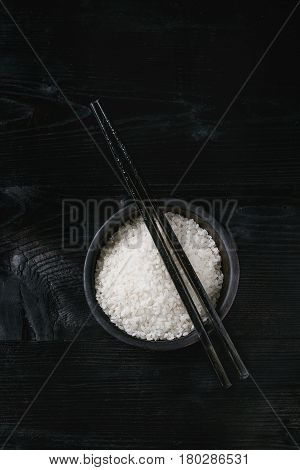 White sushi raw uncooked rice in black bowl with chopsticks over burnt wooden background. Top view with copy space