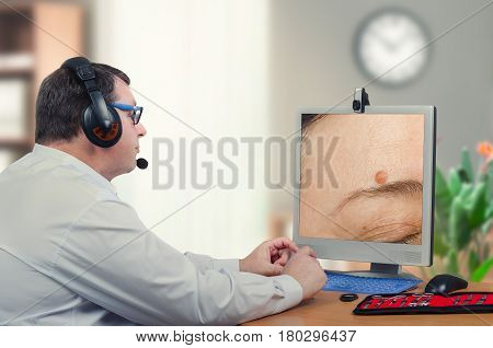 Telemedicine male dermatologist examines mole above womans eyebrow on computer monitor carefully. Virtual doctor looks at nevus on the right side of forehead either by online video chat or snapshot. Horizontal mid-shot on blurry indoors background