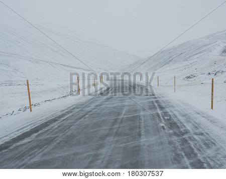 Wind Blown Snow Covered Winter Road With Mountain On The Side Of The Road Covered With Snow.