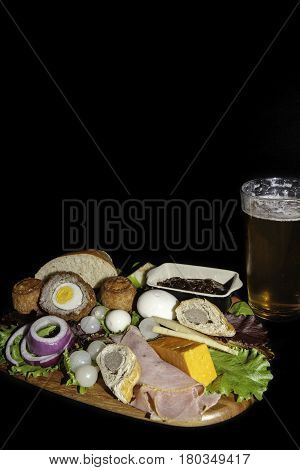 Traditional English pub grub  meal of a ploughman's lunch served with a pint of lager. The typical cold food includes pork pies ham hard boiled eggs and cheese with salad. Isolated against a black background with copy space.