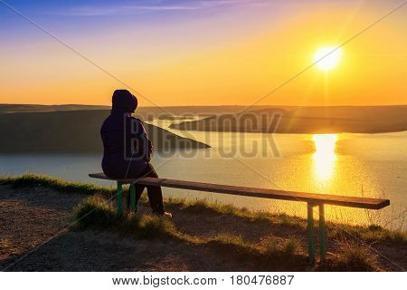 Back view of woman sitting on the bench and looking on magnificent sunset on the Dniester river sun reflected in water