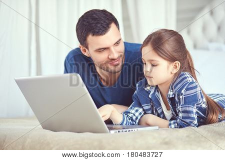 Father and his daughter are laying on the bed. Dad is watching his kid typing something on a laptop. They are both wearing pyjamas and it seems like it's morning. Education, learning, studying oncept