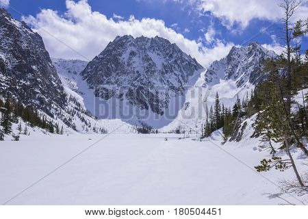 Rocky snow covered Dragontail Peak rises above frozen Colchuck Lake under bright sunshine in northern Cascade Mountains near Wenatchee Washington in April 2017