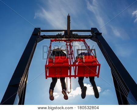Pair of people man and woman having fun in swing carousel on a high building against blue sky with backlit sunlight