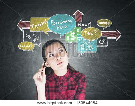 Portrait of a nerdy girl wearing a checkered shirt and sitting in a class with a colorful business plan scheme drawn on a blackboard behind her.