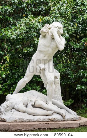 Auckland New Zealand - March 1 2017: Closeup of all white statue of slain Abel with murderer Cain standing over him and holding rock in his right hand. Green tree background.