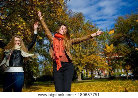 Mellow autumn. Two girls are students cheerfully spend time in the city park. Good weather blue sky. Girlfriends throw armfuls of dry yellow leaves up and rejoice as children.