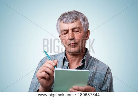 A portrait of mature man with gray hair wearing jean stylish shirt holding notebook and pen in his hands making some notes having serious expression while standing over blue background.