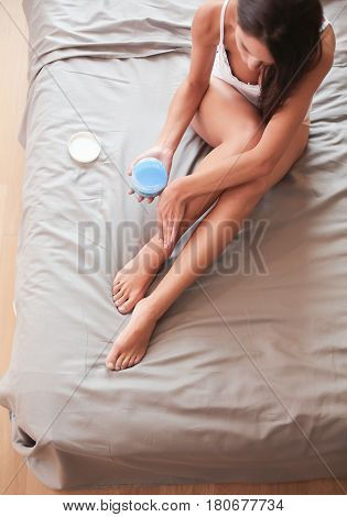 Beautiful woman sitting on bed and applying cream on legs.