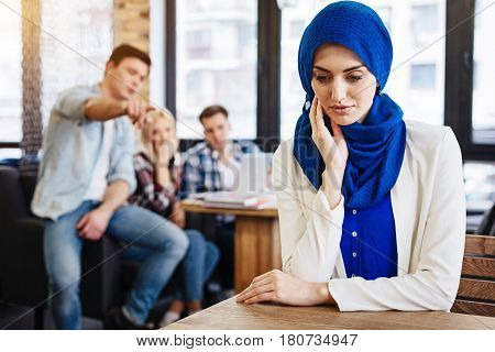 Multicultural society. Pleasant beautiful young muslim woman sitting at the table in the cafe while her mates abusing her