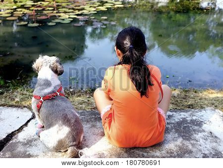 rear of little girl wearing orange t-shirts and Shih Tzu puppy with red harness sitting look natural scenic on concrete bench by the pond in public park, best friendship amidst tranquility of nature
