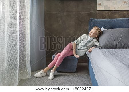 Pensive Adorable Girl Sitting On Pouffe Near The Bed At Home