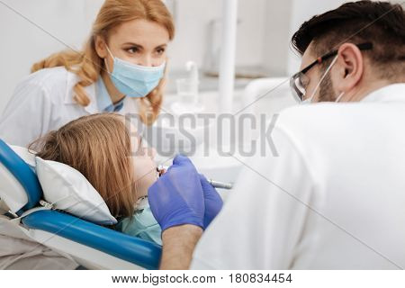 Treating baby teeth. Interest scrupulous dentist in training sitting near the doctors chair while he running a procedure using specialized equipment