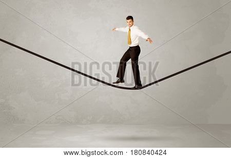 An elegan businessman in suit balancing on a tight rope with a briefcase in front of grey urban wall background environment concept
