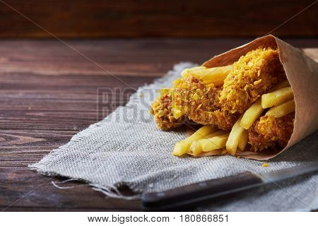 Crispy fried chicken breast with french fries to go on an old rustic wooden background with a sackcloth napkin. Close-up. With copy space.