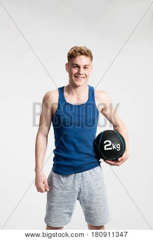 Handsome hipster fitness man in blue tank top shirt and gray shorts holding medicene ball. Studio shot on gray background.