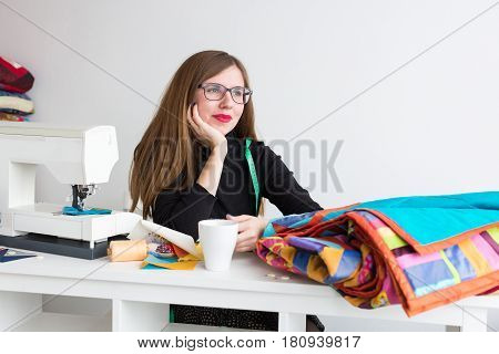 beautiful young tailor woman rests from work and looking to the side, leaning his arms on the table with a sewing machine, fabrics and cup. Quilt concept. Handcraft and hobby concept.
