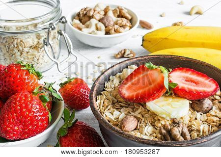 bowl of oat muesli with strawberry banana granola and nuts with ingredients for healthy breakfast on white wooden table. close up