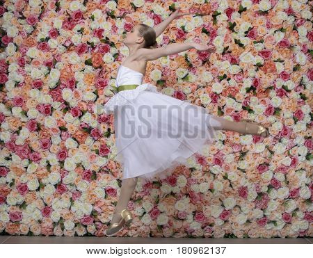 Portrait of a beautiful blonde little girl in white gown. Ballerina on a background of a flower wall in the studio