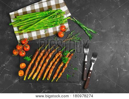 Asparagus baked grill wrapped in bacon. Ingredients for asparagus wrapped in bacon cherry tomatoes parsley thyme rosemary fresh asparagus. Dark black background. Top view from above and copy space.