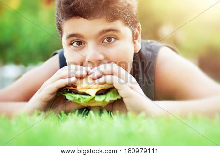 Portrait of a teen boy lying down on the grass on bright sunny day and with pleasure eating tasty burger, happy time in summer camp