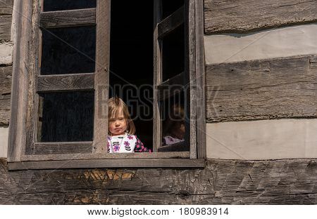 Small and lonely caucasian baby girl or toddler looking out of old wooden cabin window to suggest poverty or recession