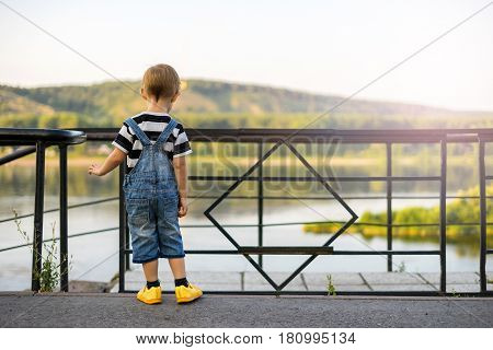 The boy stands on the edge of the ledge. The child walks along the alley off the coast. The railing closes the passage to the stairs. Cliff above the river beyond the fence.