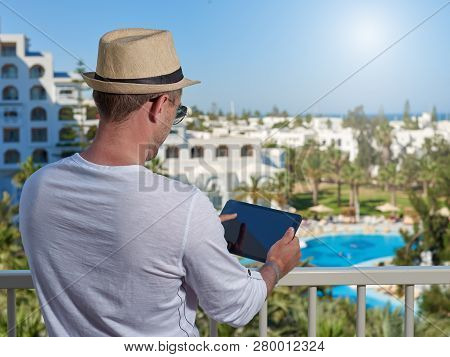 European Man In A White T-shirt And Sun Hat Is Taking Photo Of A Picteresque Hotls Veiw Using Tablet