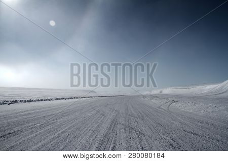 An Ice Road Leading Towards Snow Covered Hills With A Partial Sundog Located Near The Community Of C