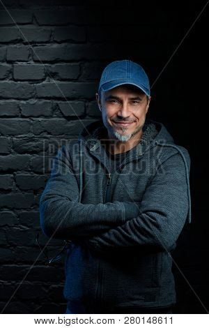 Smiling older man in jumper and baseball cap standing against black brick wall.