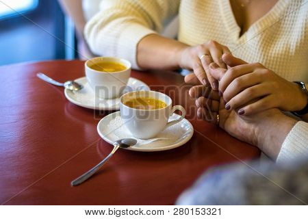 Couple Hold Hands In Cafe, Drinking Tea, Close Up Of Lovers Arms On Background Of Wooden Table With 
