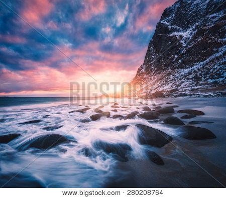 Sandy Beach With Stones In Blurred Water, Blue Sky With Pink Clouds And Snowy Mountains At Sunset. U