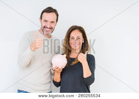 Middle age couple holding piggy bank over isolated background happy with big smile doing ok sign, thumb up with fingers, excellent sign