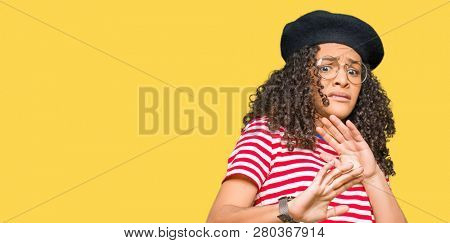 Young beautiful woman with curly hair wearing glasses and fashion beret disgusted expression, displeased and fearful doing disgust face because aversion reaction. With hands raised. Annoying concept.