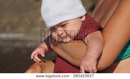 Portrait Of The Funny Baby With A Spittle In Mothers Hands Looking At Something On The Beach