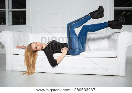 Charming Young Woman Posing Lying On The Sofa In The Studio, Dressed In A Black  T-shit And Jeans