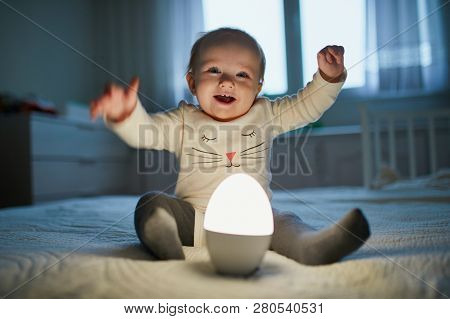 Adorable Baby Girl Playing With Bedside Lamp In Nursery. Happy Kid Sitting On Bed With Nightlight. L