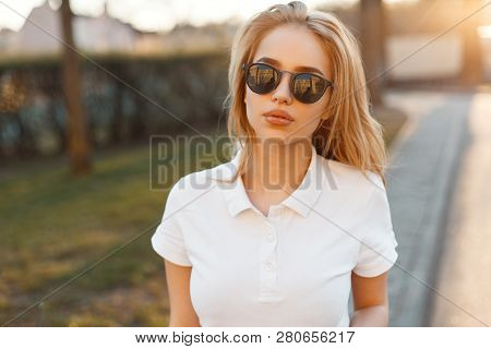 Stylish Beautiful Young Hipster Woman In Trendy Sunglasses In A White Polo T-shirt Is Standing On Th