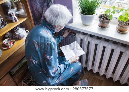 The Senior Woman Holding Gas Bill In Front Of Heating Radiator. Payment For Heating In Winter.