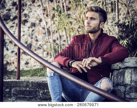 Handsome Young Man Outdoor In Winter Fashion, Wearing Red Jacket And Woolen Scarf In City Park