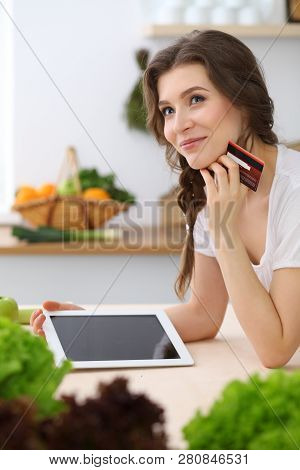 Young  Woman Looking For A New Recipe For  Cooking In A Kitchen. Housewife Is Making Online Shopping