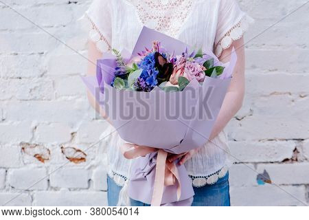A Bouquet Of Flowers, Packed In Purple Paper In The Hands Of A Woman On A Light Background. Bouquet 