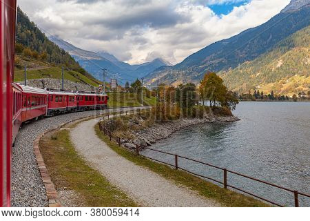 Beautiful View Of  Red  Rhaetian Train Running On The Lake Side Of Lago Di Poschiavo In Autumn With 