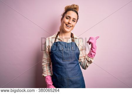 Young beautiful blonde cleaner woman doing housework wearing arpon and gloves smiling with happy face looking and pointing to the side with thumb up.