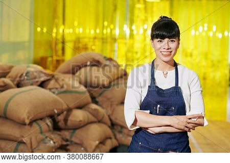 Portrait Of Pretty Smiling Female Roastery Owner In Denim Aprom Standing In Warehouse In Front Of Co