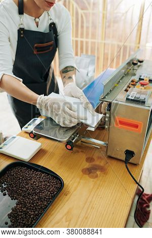 Female Roastery Worker In Apron And Rubber Gloves Sealing Seams Of Coffee Packages